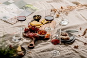 Ein stilvolles Picknick-Arrangement mit einem Becher-Set, bestehend aus Weingläsern und Wassergläsern, auf einer Decke. Im Vordergrund sind Käse, Trauben, Salami und Brot auf einem Holzbrett zu sehen. Perspektive von oben.
