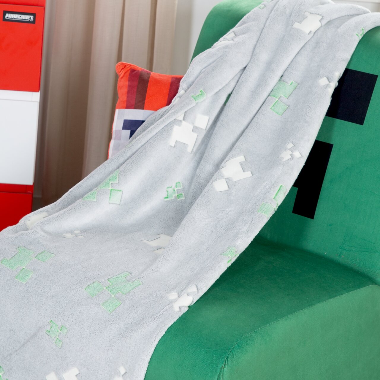 Een lichtgrijze, zachte Minecraft-deken met groene en witte pixelmotieven, gedrapeerd over een groene Creeper-fauteuil, schuin van voren gefotografeerd.