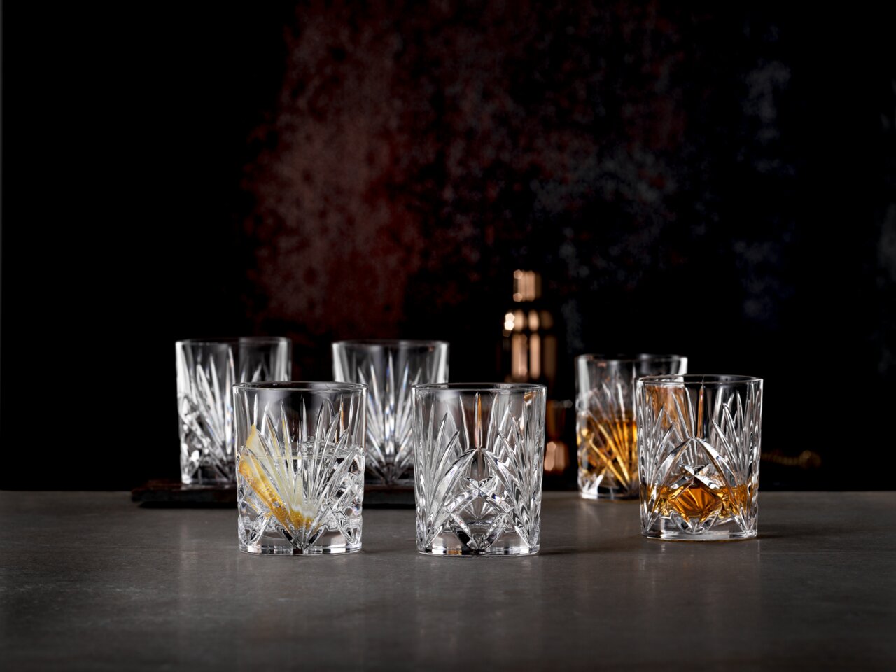 Sechs elegante Whiskybecher aus Kristallglas mit feinem Schliffmuster, frontal auf einem Tisch arrangiert, im Hintergrund ein Cocktailshaker.