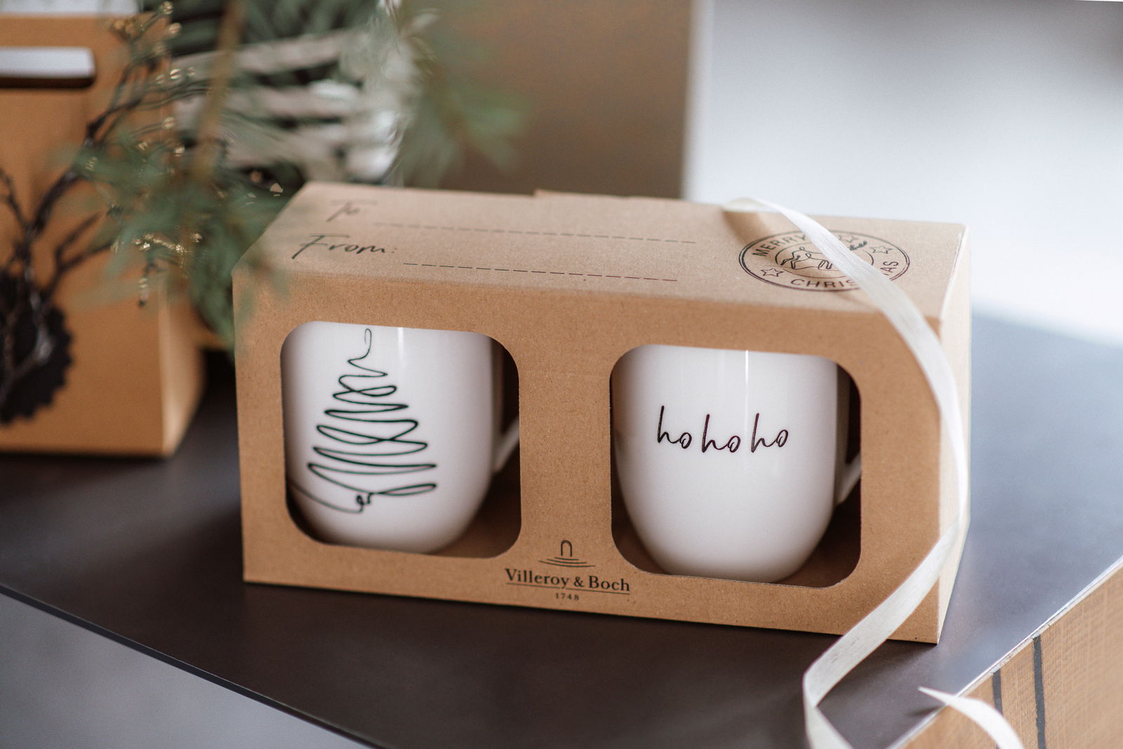 Zwei weiße Tassen in einer Geschenkverpackung mit Weihnachtsmotiven, eine mit einem stilisierten Baum und die andere mit 'ho ho ho' Schriftzug, seitliche Perspektive.