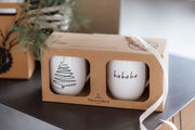 Zwei weiße Tassen in einer Geschenkverpackung mit Weihnachtsmotiven, eine mit einem stilisierten Baum und die andere mit 'ho ho ho' Schriftzug, seitliche Perspektive.
