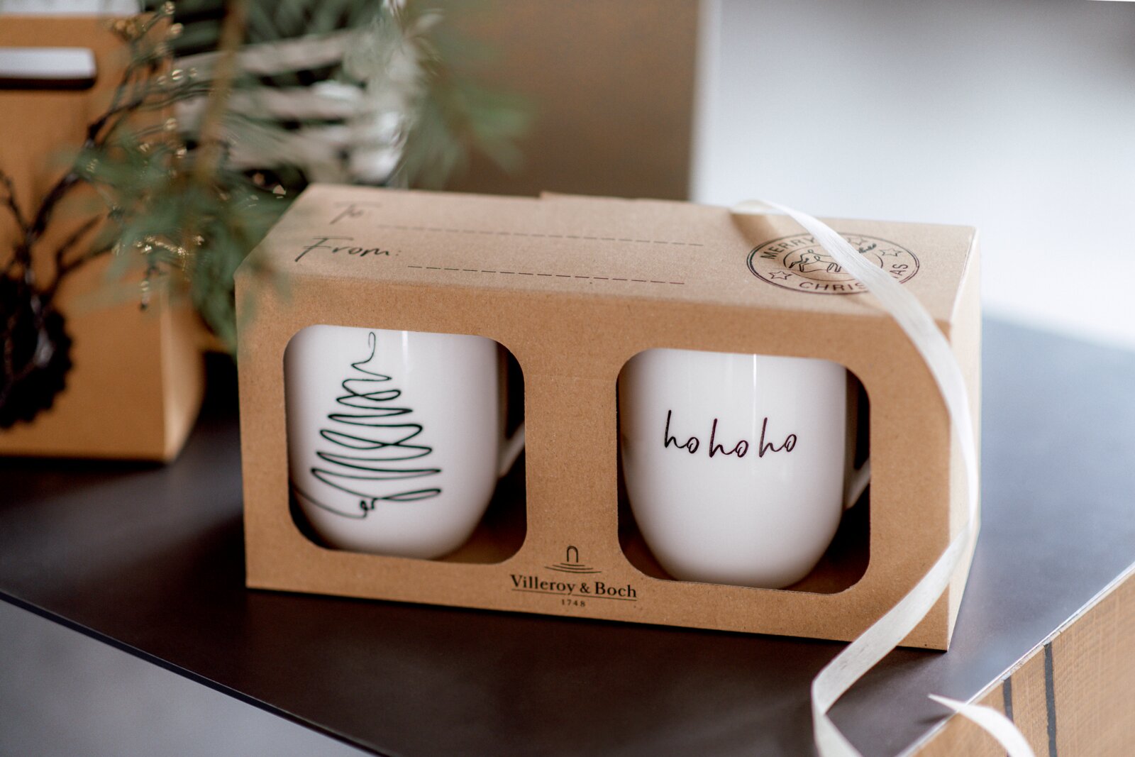 Zwei weiße Tassen in einer Geschenkverpackung mit Weihnachtsmotiven, eine mit einem stilisierten Baum und die andere mit 'ho ho ho' Schriftzug, seitliche Perspektive.
