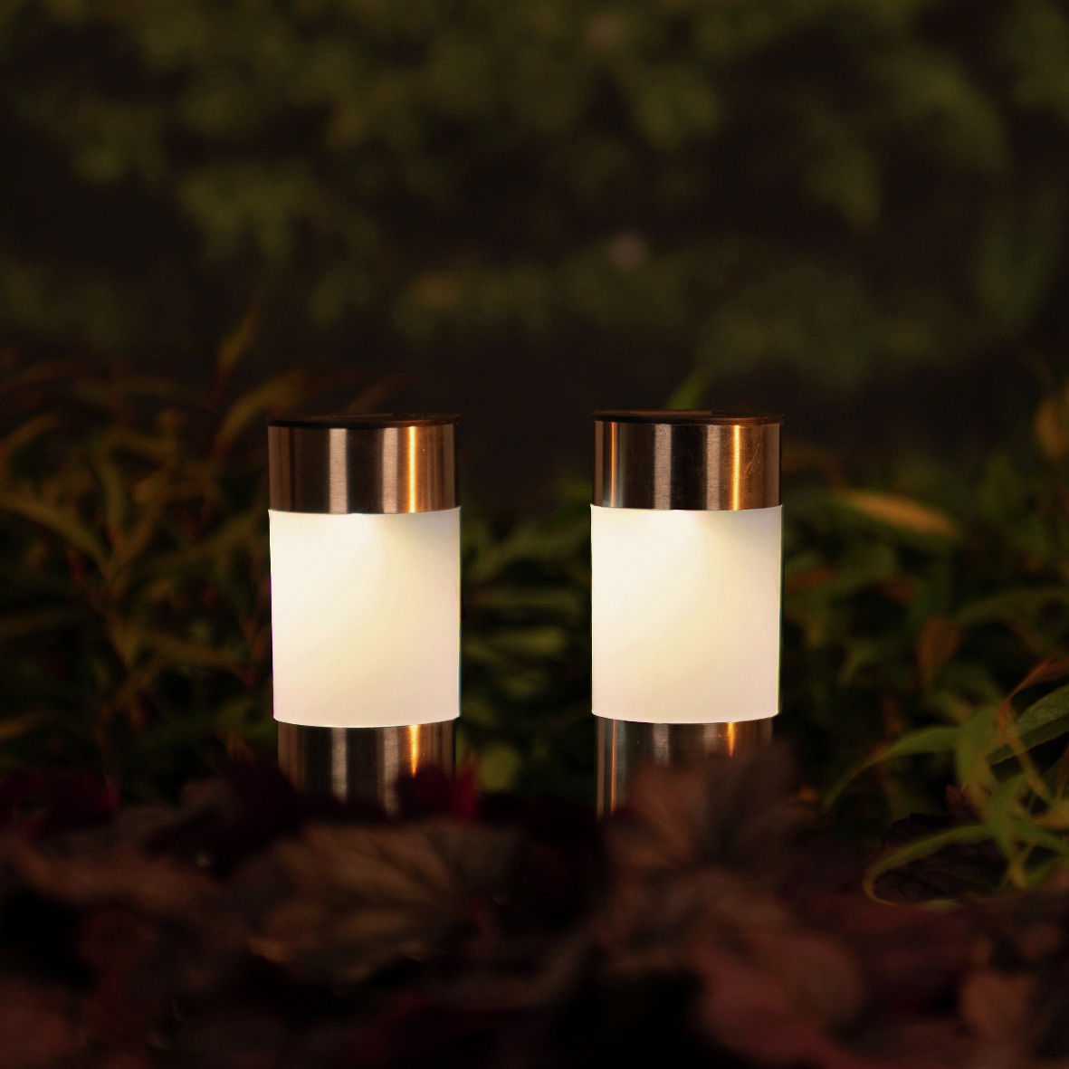 Zwei Solar Stabbeleuchten aus Edelstahl in einem Garten, frontal fotografiert, umgeben von Pflanzen.