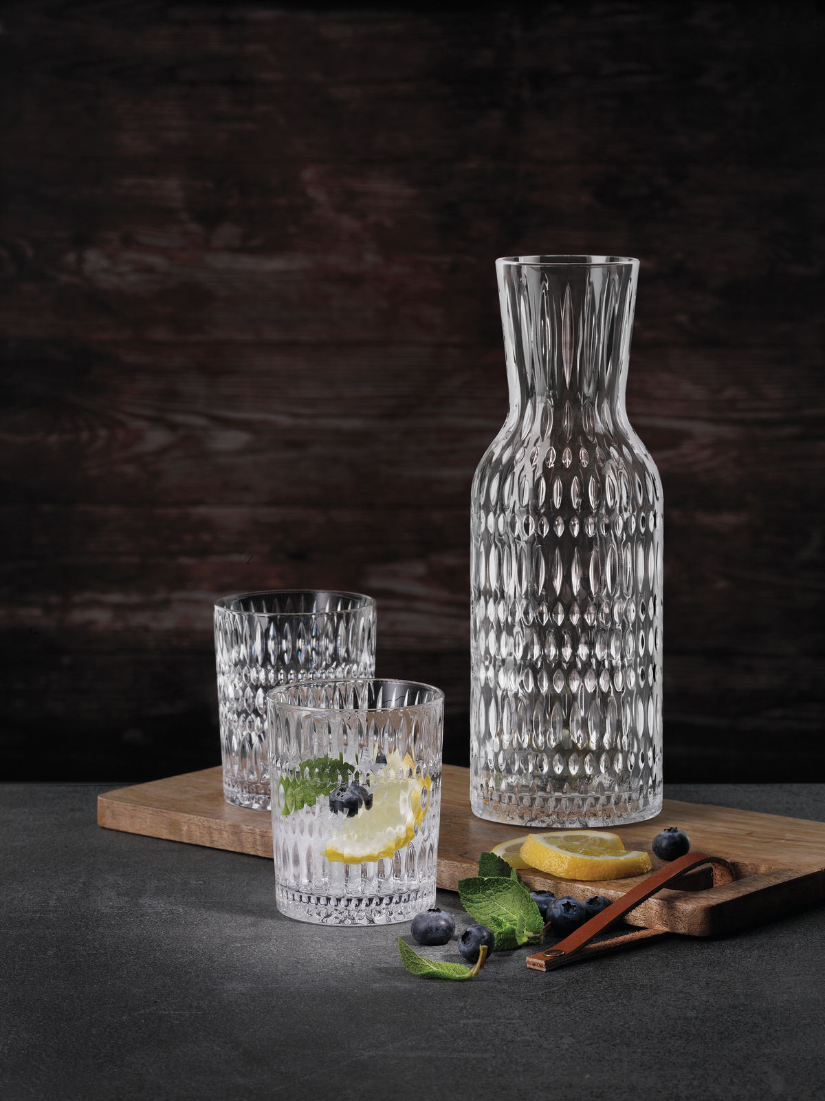 Nachtmann Trinkbecher Set 3-tlg. ETHNO Set aus einer eleganten Glaskaraffe und zwei passenden Trinkgläsern mit strukturierter Oberfläche, präsentiert auf einem Holzbrett mit Zitronenscheiben und Blaubeeren, seitliche Perspektive.