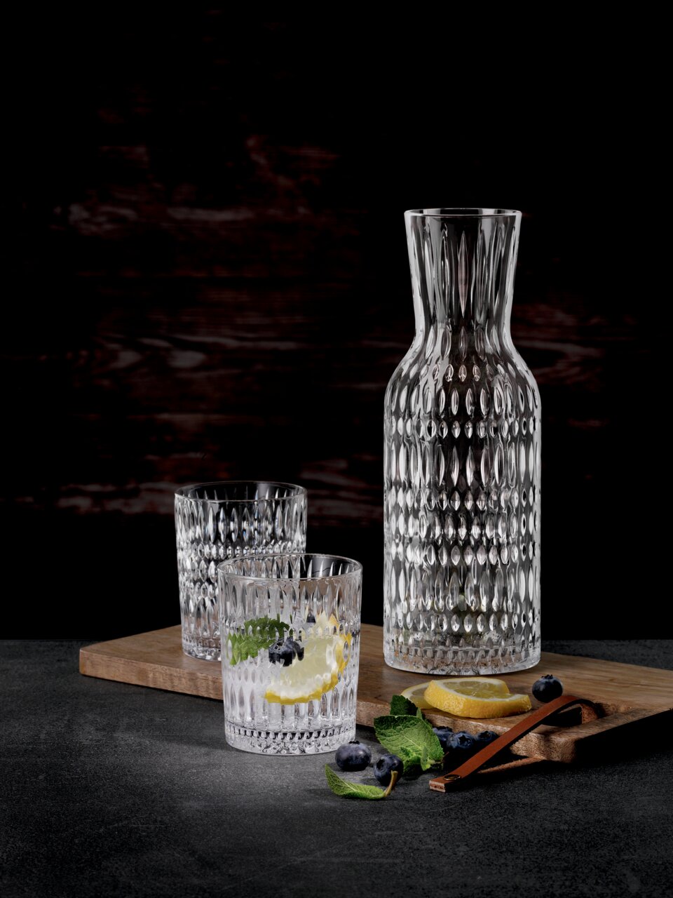Nachtmann Trinkbecher Set 3-tlg. ETHNO Set aus einer eleganten Glaskaraffe und zwei passenden Trinkgläsern mit strukturierter Oberfläche, präsentiert auf einem Holzbrett mit Zitronenscheiben und Blaubeeren, seitliche Perspektive.