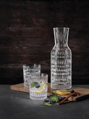 Set aus einer eleganten Glaskaraffe und zwei passenden Trinkgläsern mit strukturierter Oberfläche, präsentiert auf einem Holzbrett mit Zitronenscheiben und Blaubeeren, seitliche Perspektive.