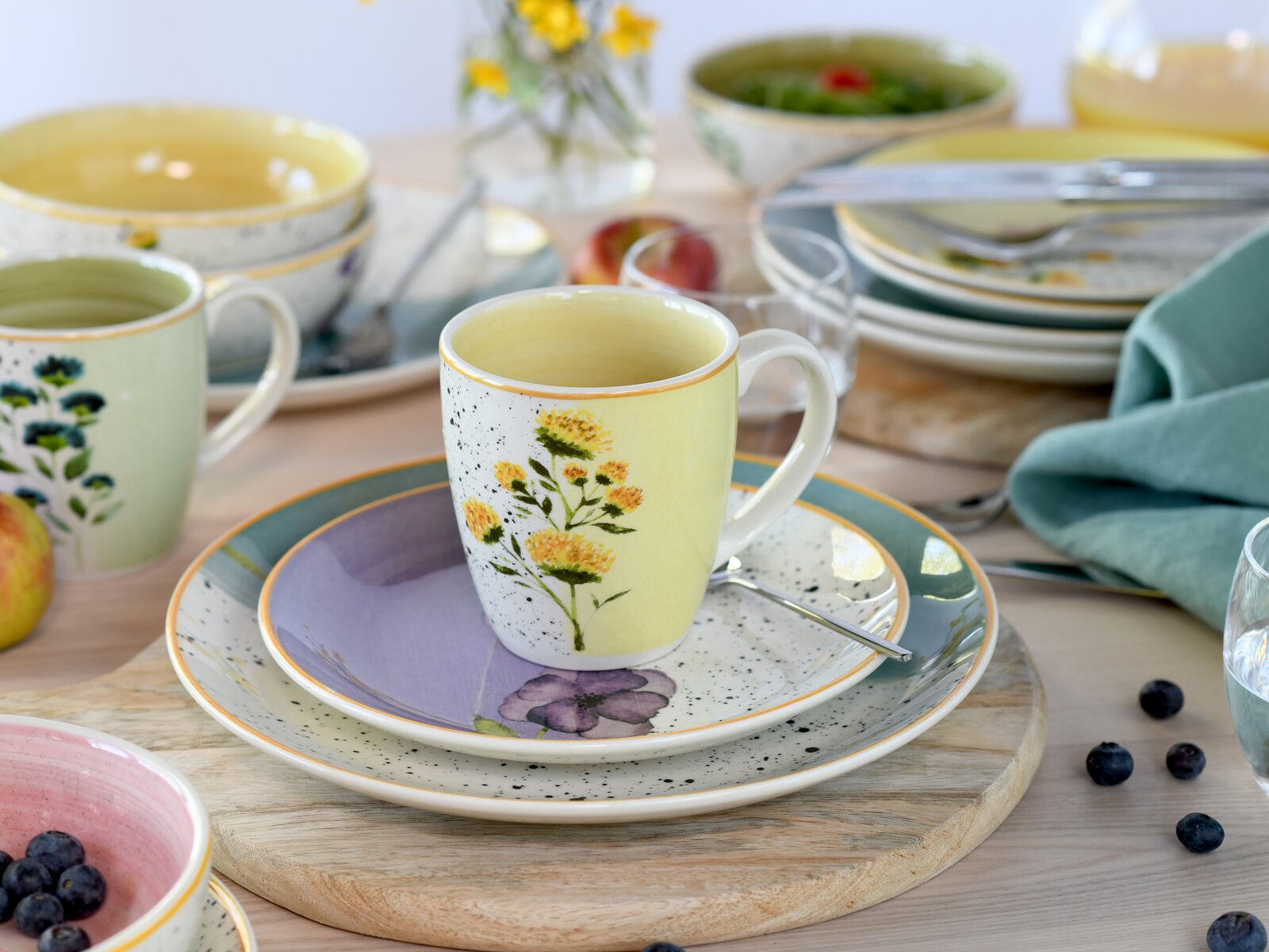 Leicht von oben fotografierte gelb-weiße Tasse mit floralem Motiv auf farbenfrohen, gesprenkelten Tellern in einer sommerlich gedeckten Tischszene