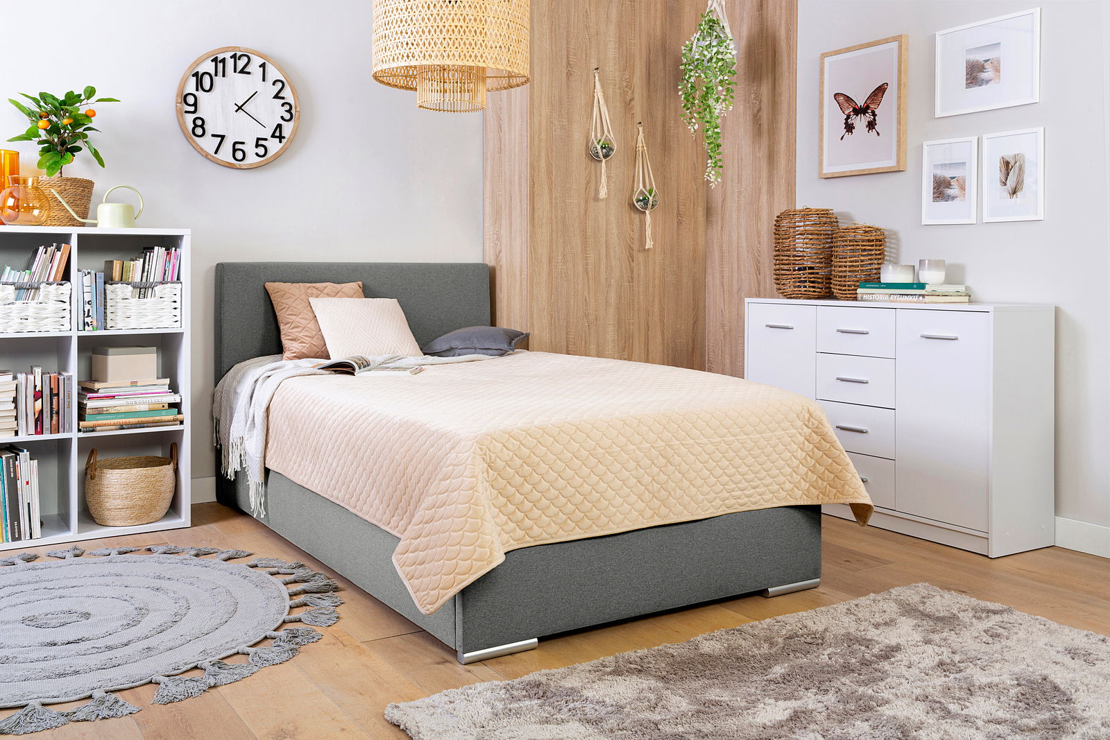 Gemütliches Schlafzimmer mit einem grauen Boxbett, dekoriert mit Kissen und einer beigen Steppdecke, umgeben von modernen Möbeln und Pflanzen. Perspektive von der Seite.