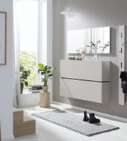 Wandhängender weißer Schuhschrank mit zwei grifflosen Klappen, aus leicht seitlicher Frontperspektive in einem hellen modernen Flur gezeigt.