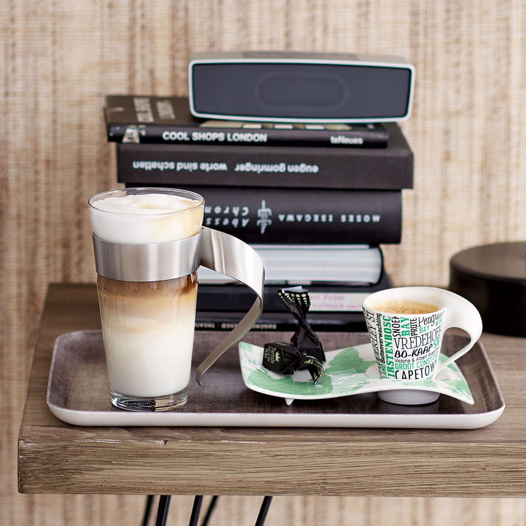 Latte Macchiato Glas 0,3l mit Edelstahlgriff, gefüllt mit geschichtetem Kaffee und Milchschaum, auf einem Tablett neben einer Espressotasse, seitliche Perspektive