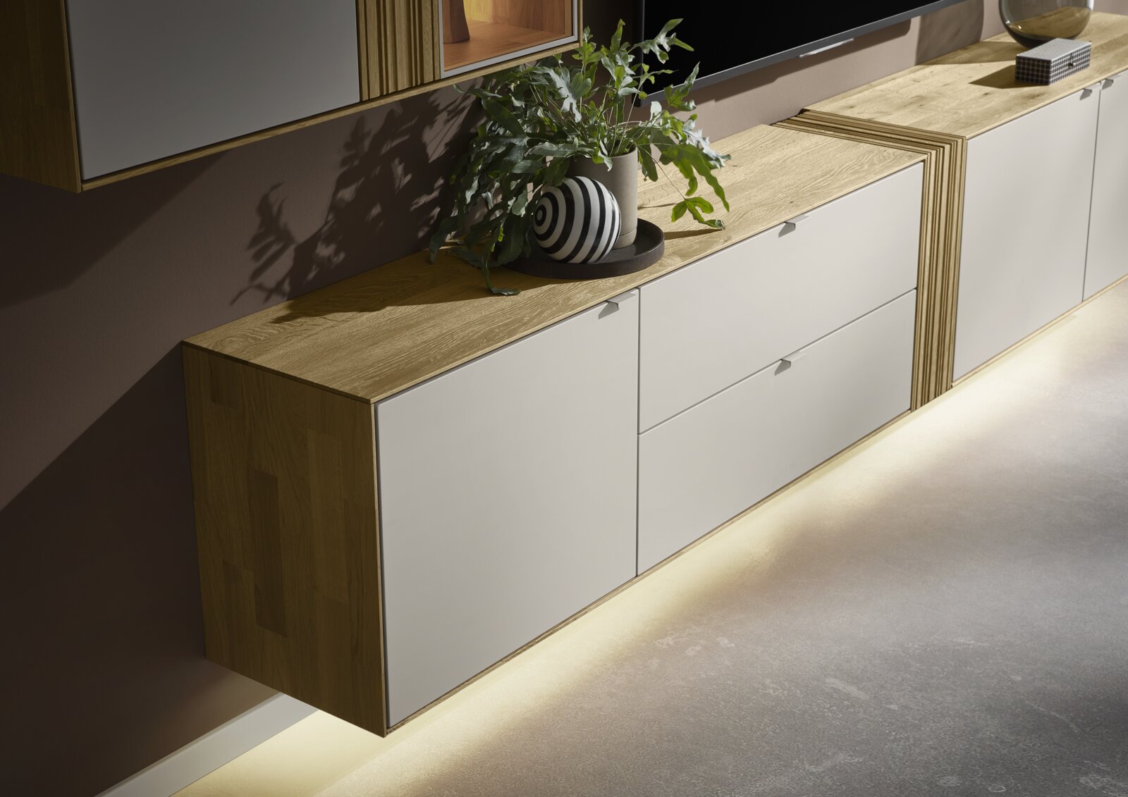 Schuin vooraanzicht van een modern zwevend tv-meubel/dressoir met eikenhouten blad en zijkanten, matte witte fronten en warme indirecte LED-verlichting onderaan, gestyled met plant en decoratieve vaas.
