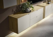 Schuin vooraanzicht van een modern zwevend tv-meubel/dressoir met eikenhouten blad en zijkanten, matte witte fronten en warme indirecte LED-verlichting onderaan, gestyled met plant en decoratieve vaas.