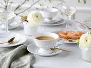 Elegantes Kaffeegedeck aus weißem Porzellan mit goldfarbenem Besteck, Espresso in Tasse, Gebäck und weißen Blumen auf heller Tischdecke, fotografiert in leichter Schrägansicht von vorne.