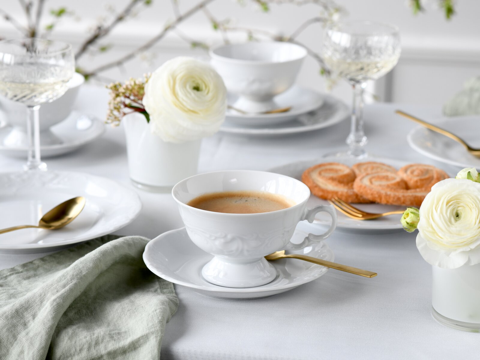Elegantes Kaffeegedeck aus weißem Porzellan mit goldfarbenem Besteck, Espresso in Tasse, Gebäck und weißen Blumen auf heller Tischdecke, fotografiert in leichter Schrägansicht von vorne.