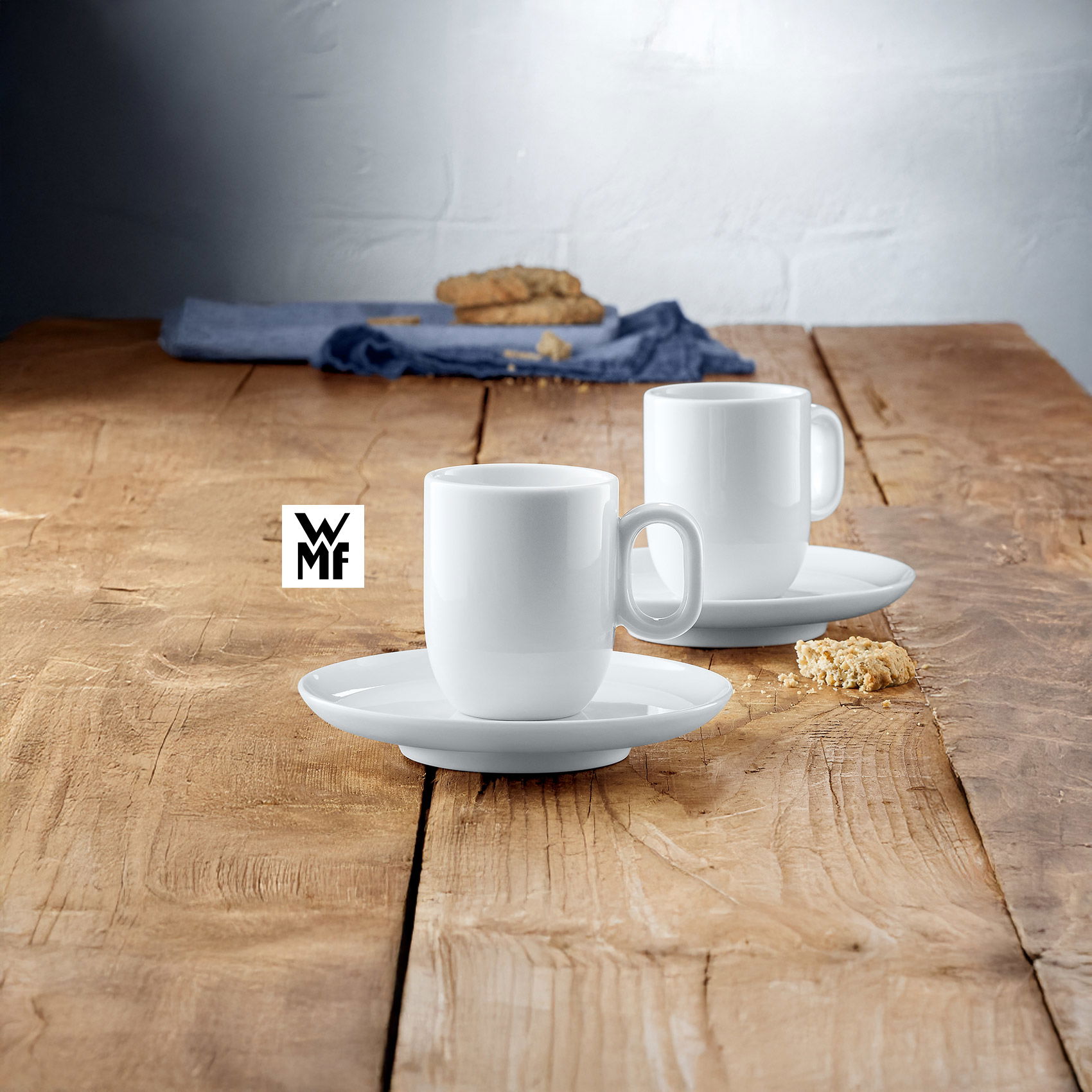 Zwei weiße WMF-Kaffeetassen mit Untertellern auf rustikalem Holztisch, umgeben von Kekskrümeln und Stoffserviette