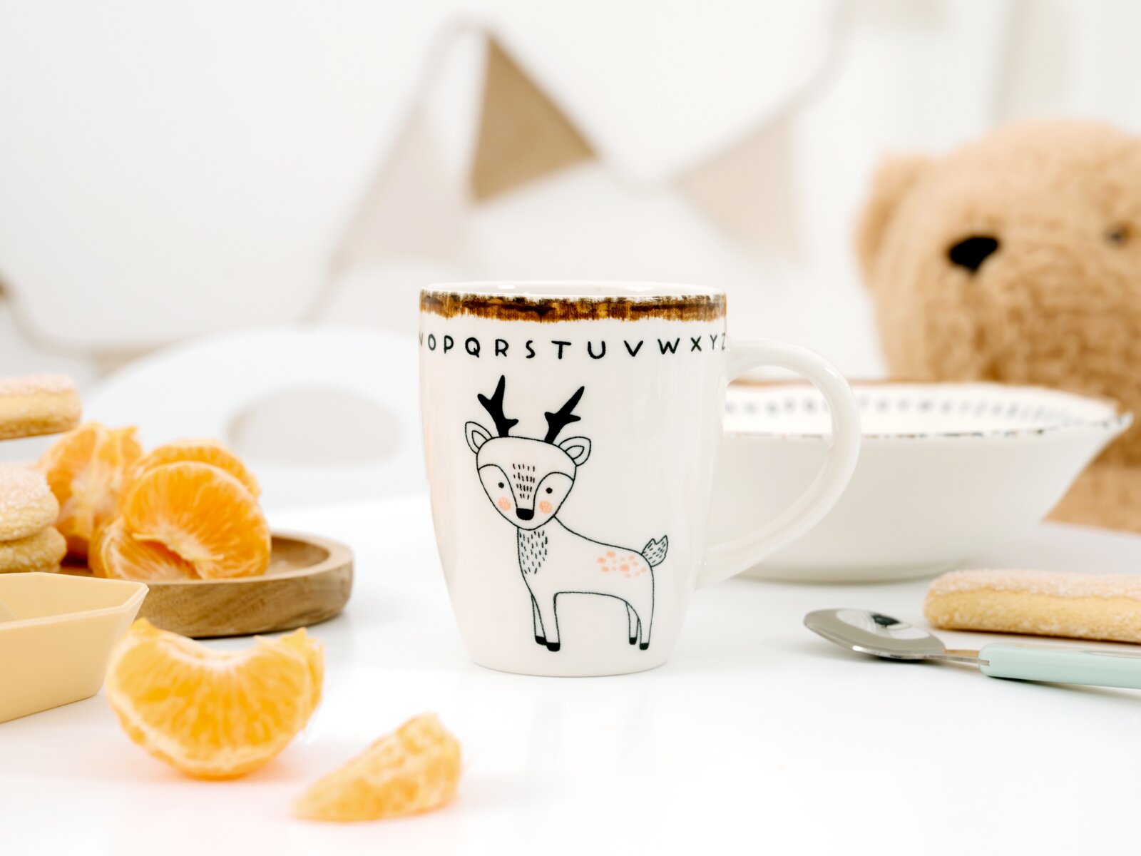 Weiße Kindertasse mit niedlichem Hirschmotiv und Buchstabenrand, frontal aus leicht erhöhter Perspektive in einer hellen Frühstücksszene mit Mandarinen und Teddybär