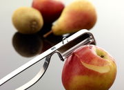 Nahaufnahme eines Pendel-Sparschälers, der eine rote Apfelschale entfernt. Im Hintergrund sind unscharf eine Birne und eine Kiwi zu sehen.