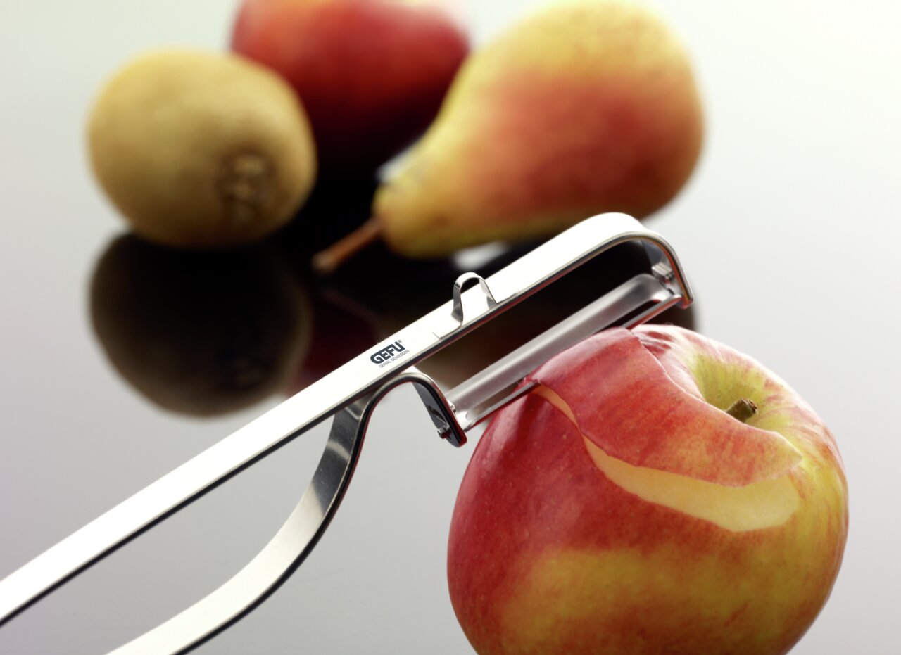 Nahaufnahme eines Pendel-Sparschälers, der eine rote Apfelschale entfernt. Im Hintergrund sind unscharf eine Birne und eine Kiwi zu sehen.