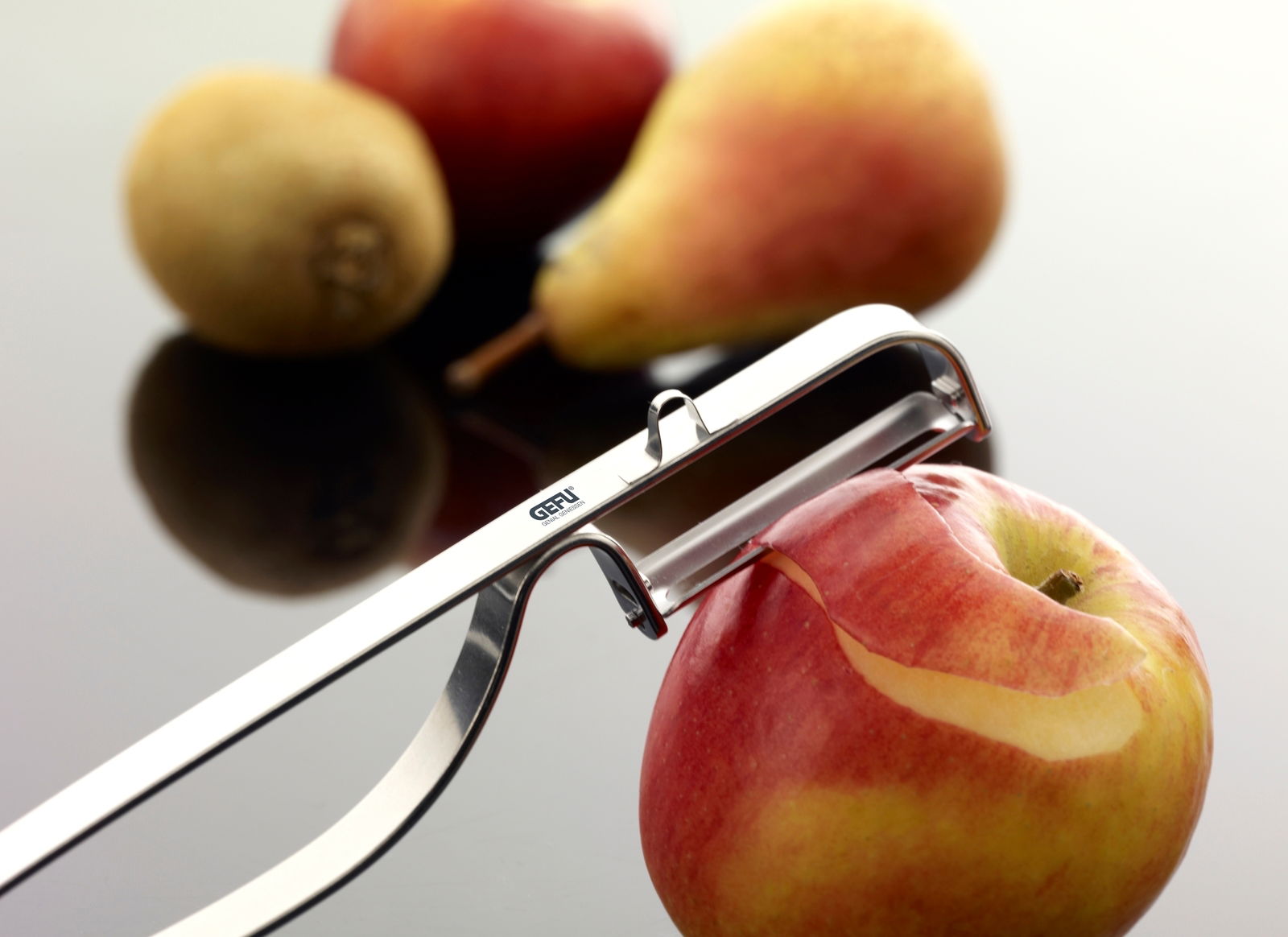 Nahaufnahme eines Pendel-Sparschälers, der eine rote Apfelschale entfernt. Im Hintergrund sind unscharf eine Birne und eine Kiwi zu sehen.