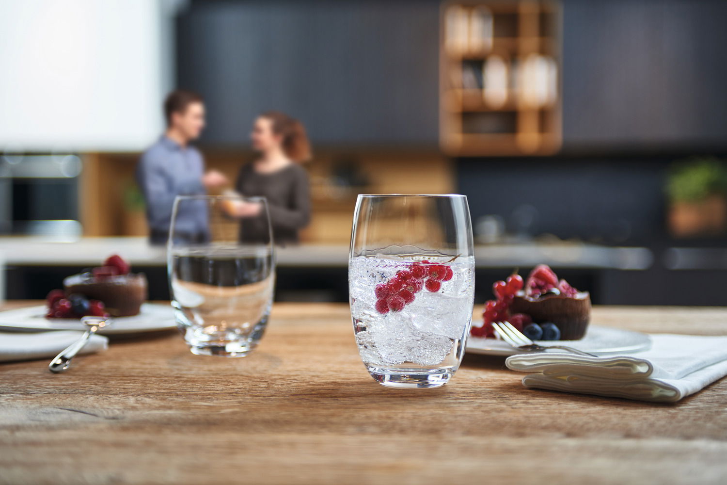 Trinkglas Cheers 460ml mit Eiswürfeln und roten Beeren, auf einem Holztisch in einer modernen Küche, seitliche Perspektive.
