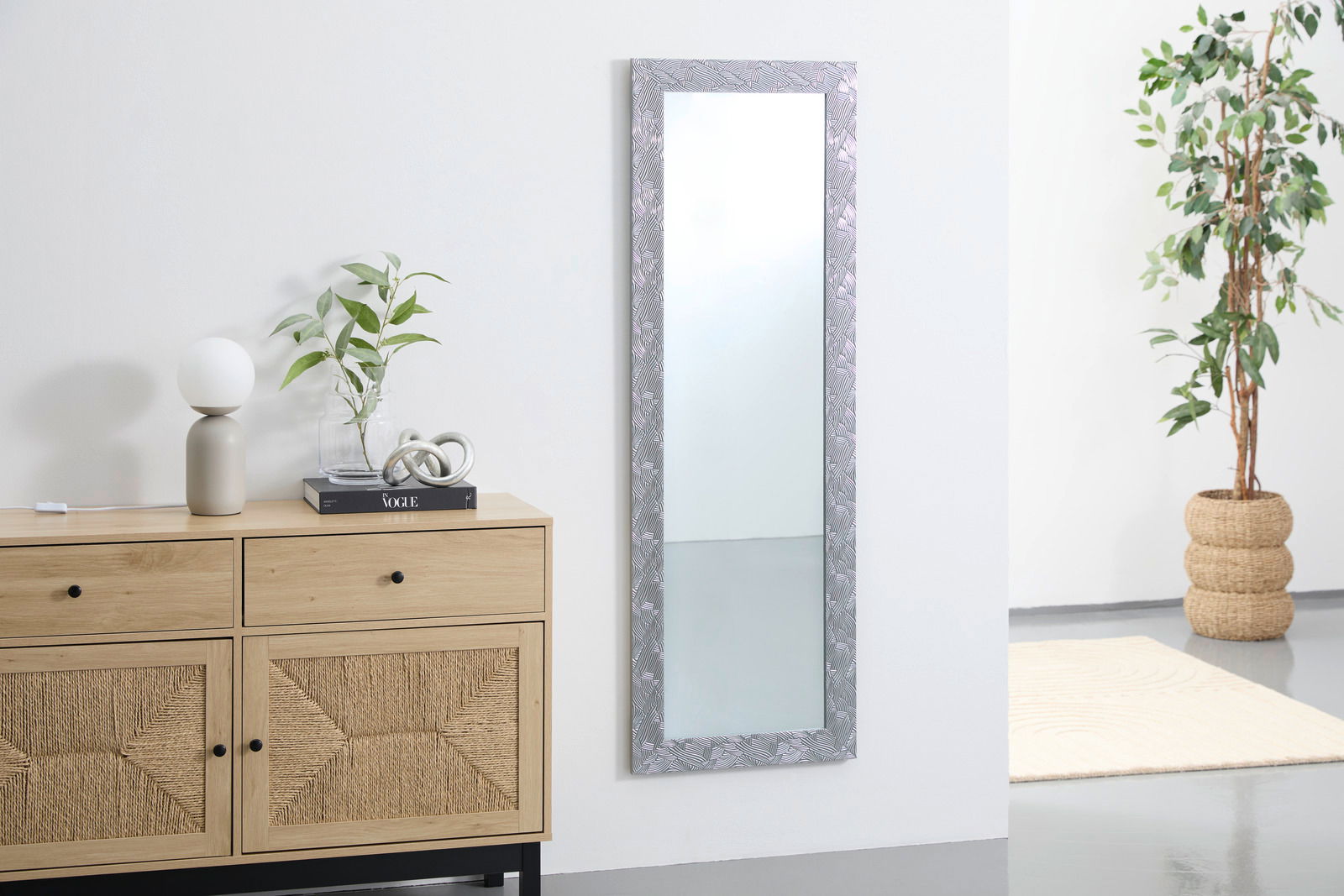 Rechteckiger Spiegel mit dekorativem Rahmen, an einer weißen Wand hängend, neben einer Kommode und einer Pflanze, aus seitlicher Perspektive.