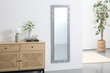 Rechteckiger Spiegel mit dekorativem Rahmen, an einer weißen Wand hängend, neben einer Kommode und einer Pflanze, aus seitlicher Perspektive.