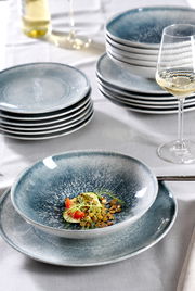 Elegantes Tellerset 18-tlg in Blau-Grau, gestapelt auf einem Tisch mit einem gefüllten Teller im Vordergrund, seitliche Perspektive.