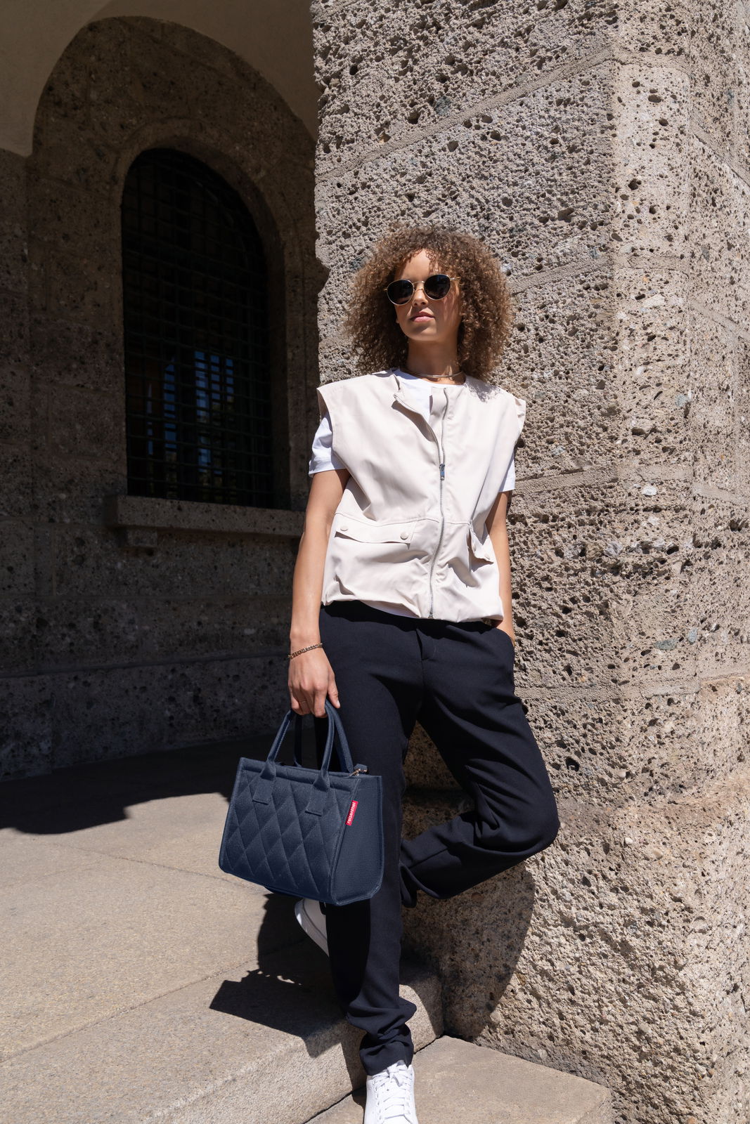 Frau mit Sonnenbrille lehnt an einer Steinwand und hält eine kleine, gesteppte, dunkelblaue Tasche in der Hand. Die Perspektive ist von vorne.