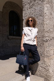 Frau mit Sonnenbrille lehnt an einer Steinwand und hält eine kleine, gesteppte, dunkelblaue Tasche in der Hand. Die Perspektive ist von vorne.