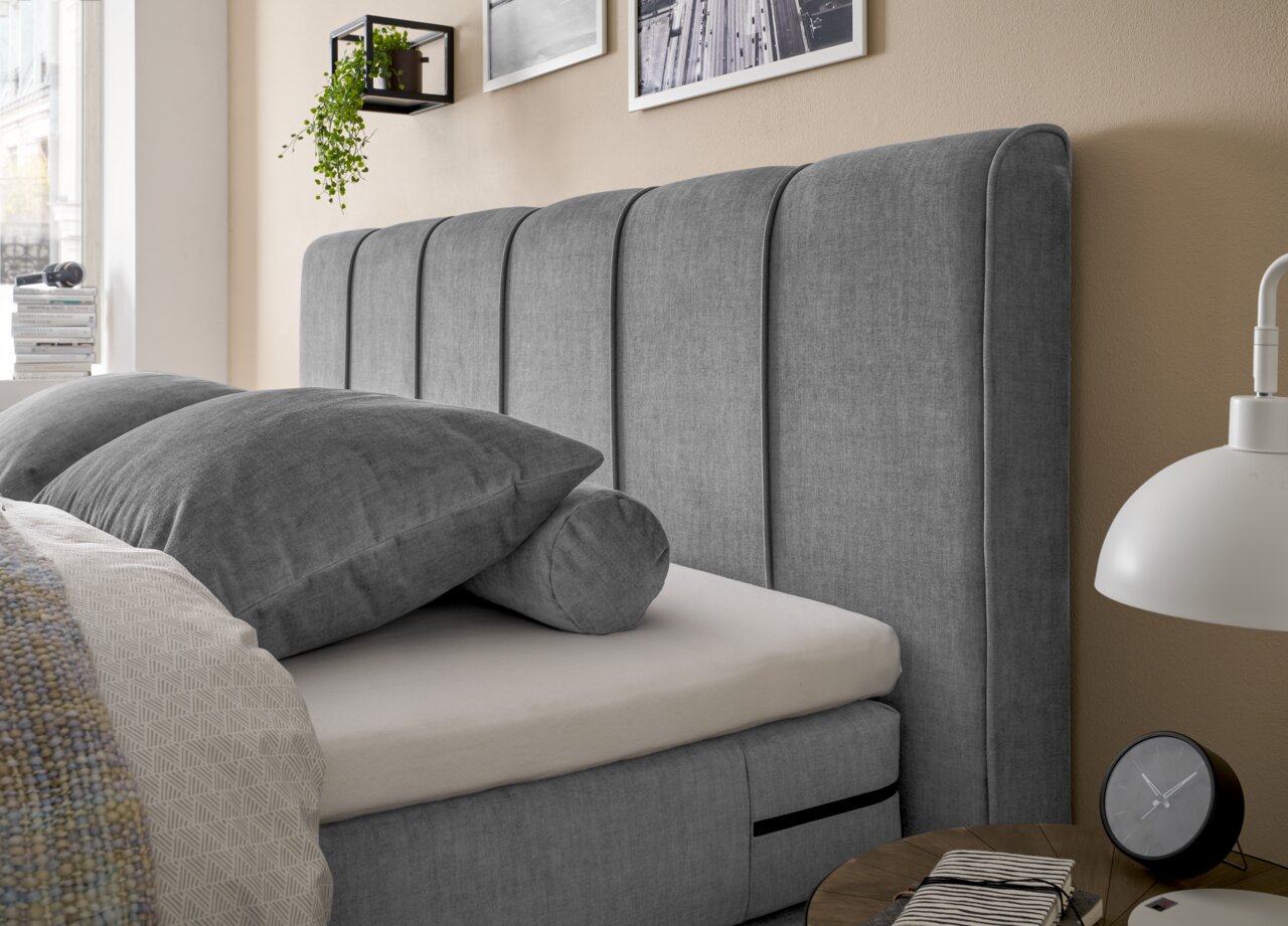 Set one by Musterring Boxspringbett SET ONE FAIRFIELD Seitliche Perspektive eines grauen Boxspringbetts mit gepolstertem Kopfteil und dekorativen Kissen.
