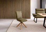 Olivgrüner Polsterstuhl mit Holz-Drehgestell im modernen Wohnzimmer, frontale Ansicht in leichter Seitenperspektive vor Holzlamellenwand und neben Couchtisch