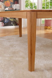 Nahaufnahme eines Esstisches aus Holz mit robusten Beinen, auf einem weißen Teppich stehend, mit einem dekorativen Hintergrund aus Blumen und einem Fenster mit Blick ins Grüne.