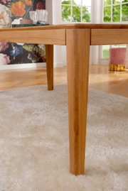 Nahaufnahme eines Esstisches aus Holz mit robusten Beinen, auf einem weißen Teppich stehend, mit einem dekorativen Hintergrund aus Blumen und einem Fenster mit Blick ins Grüne.