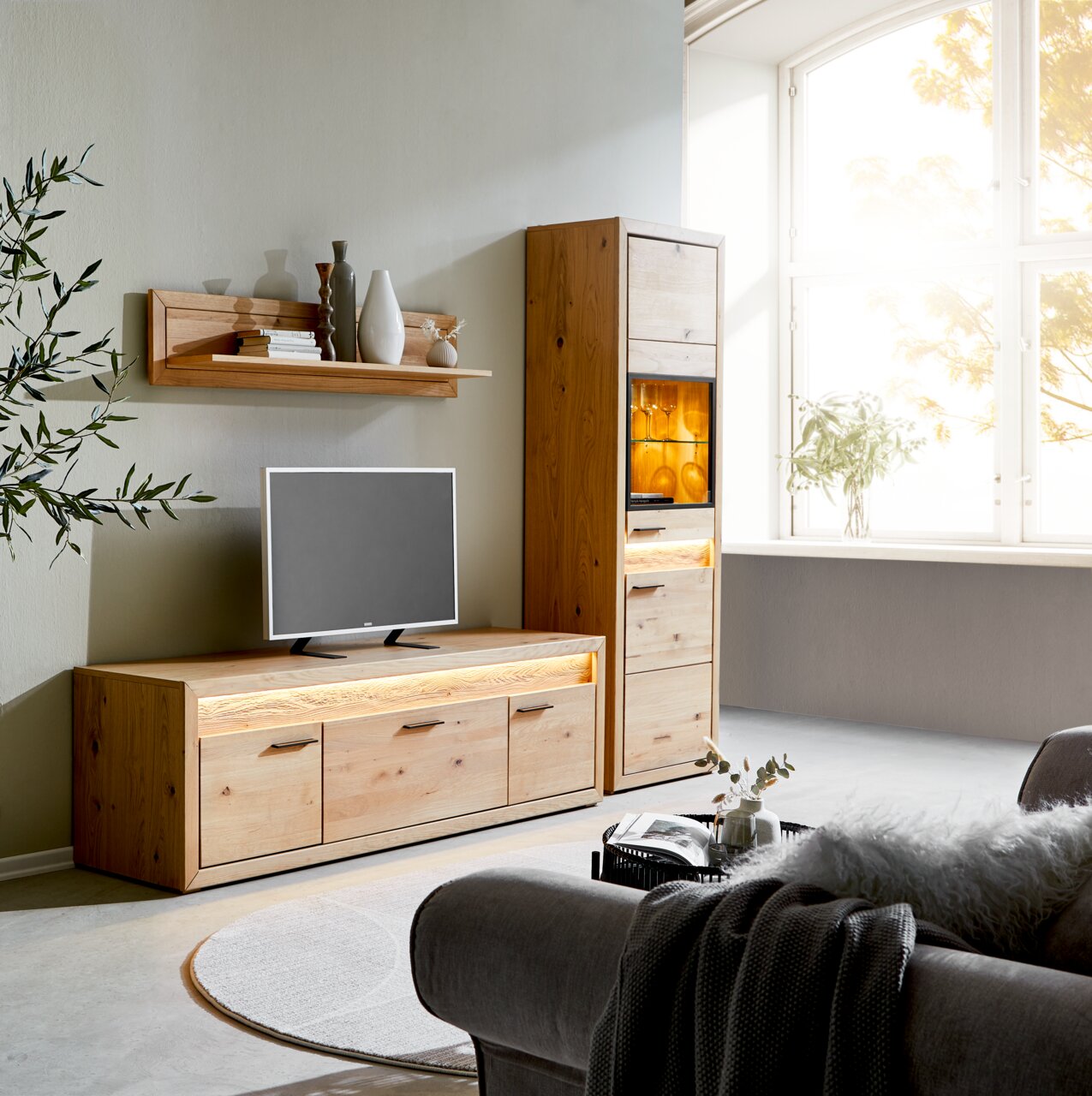 Zicht op de woonkamer met Cantus-vitrine CLEVELAND, lowboard en wandplank van hout, verlicht, met decoraties en tv, zijaanzicht.