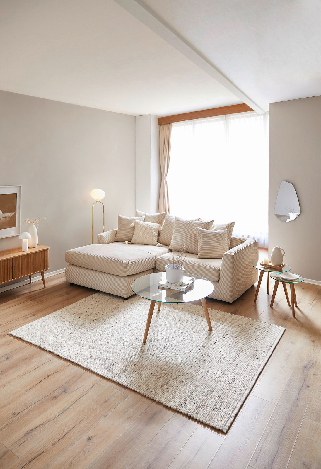 Helles Wohnzimmer mit einer beigen Rundecke, die von der Seite betrachtet wird. Die Rundecke ist mit mehreren Kissen dekoriert und steht auf einem hellen Holzboden. Ein runder Glastisch und ein Teppich ergänzen das gemütliche Ambiente.