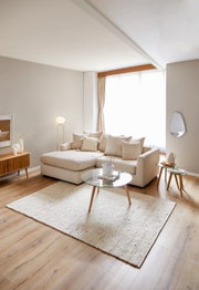 Helles Wohnzimmer mit einer beigen Rundecke, die von der Seite betrachtet wird. Die Rundecke ist mit mehreren Kissen dekoriert und steht auf einem hellen Holzboden. Ein runder Glastisch und ein Teppich ergänzen das gemütliche Ambiente.