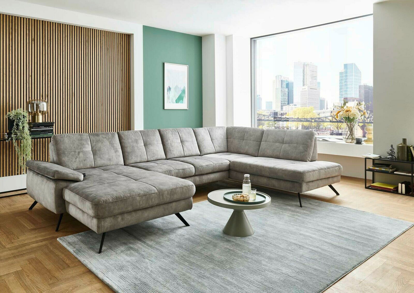 Graue Polsterrundecke in modernem Wohnzimmer mit Blick auf die Stadt durch ein großes Fenster, aus seitlicher Perspektive fotografiert.