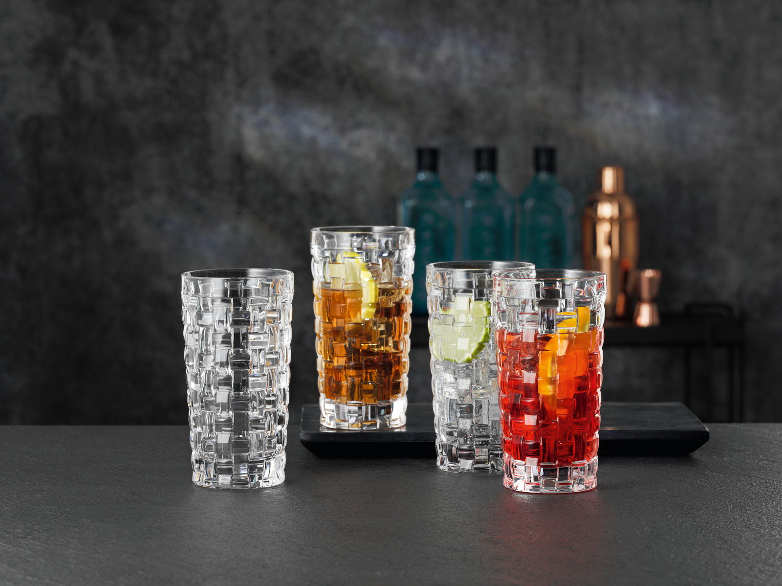 Drei Longdrinkgläser Bossa Nova mit verschiedenen Getränken und Eiswürfeln, von vorne betrachtet, auf einem dunklen Tisch vor einem unscharfen Hintergrund.