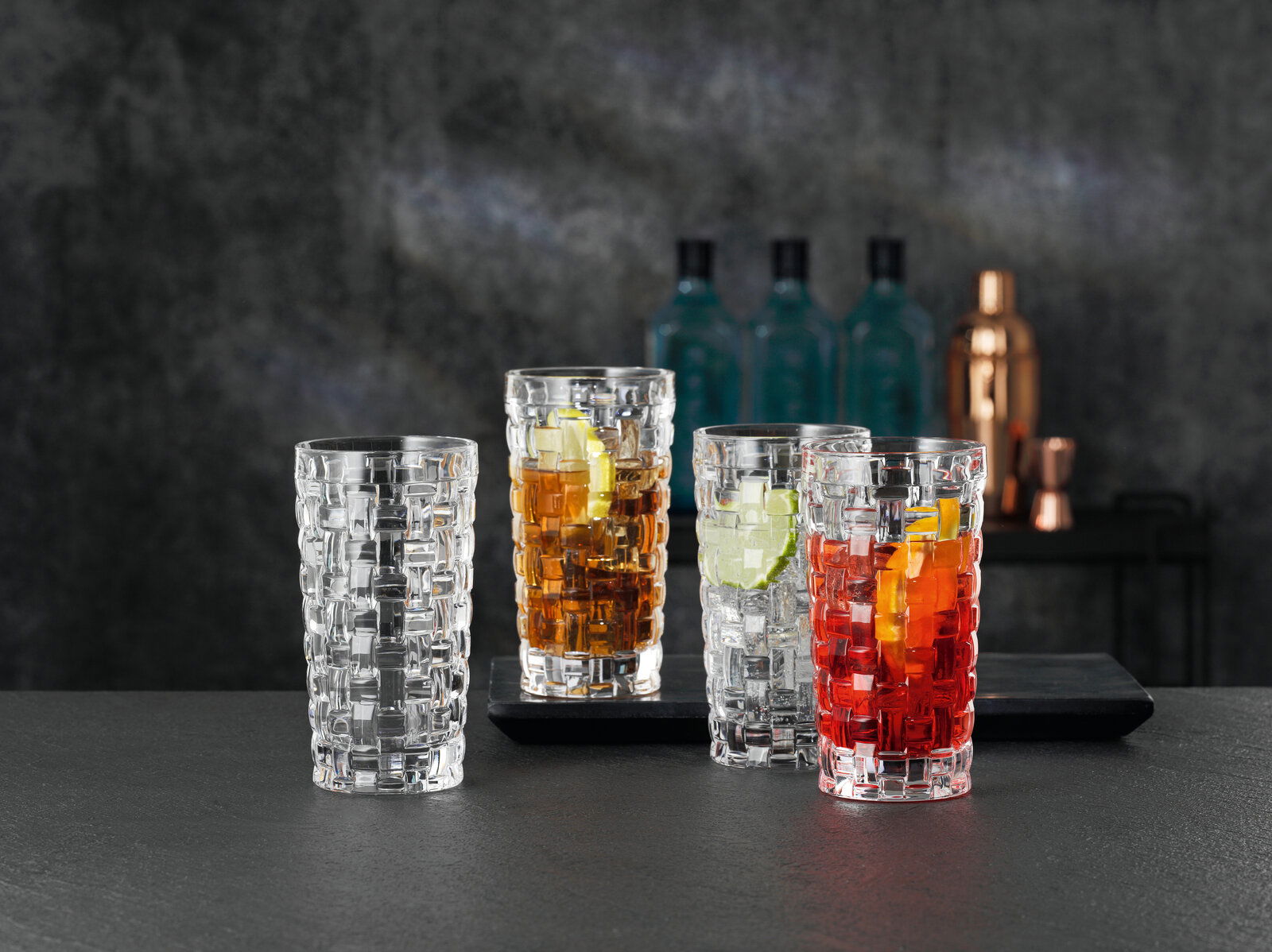 Nachtmann Longdrinkglas BOSSA NOVA Drei Longdrinkgläser Bossa Nova mit verschiedenen Getränken und Eiswürfeln, von vorne betrachtet, auf einem dunklen Tisch vor einem unscharfen Hintergrund.