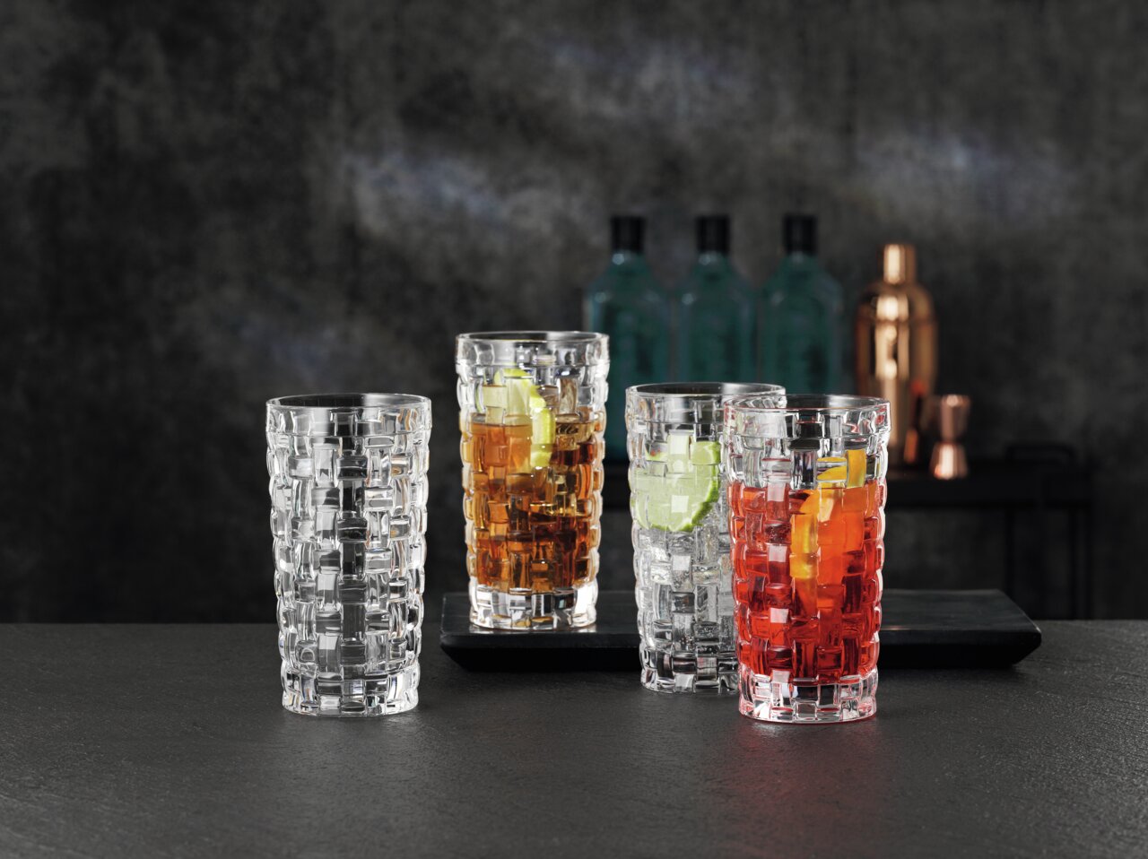 Nachtmann Longdrinkglas BOSSA NOVA Drei Longdrinkgläser Bossa Nova mit verschiedenen Getränken und Eiswürfeln, von vorne betrachtet, auf einem dunklen Tisch vor einem unscharfen Hintergrund.