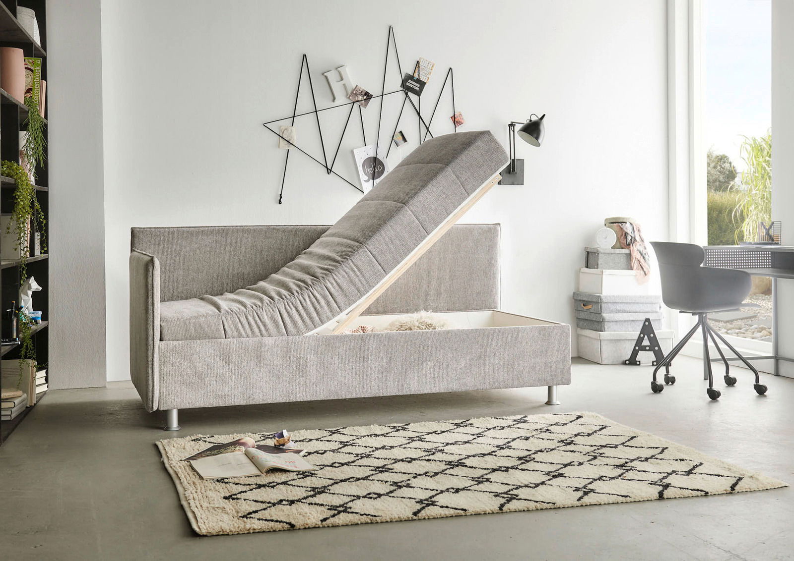 Graue Polsterliege mit hochgeklappter Matratze, seitliche Perspektive, zeigt Stauraum darunter, modernes Design in einem hellen Raum.