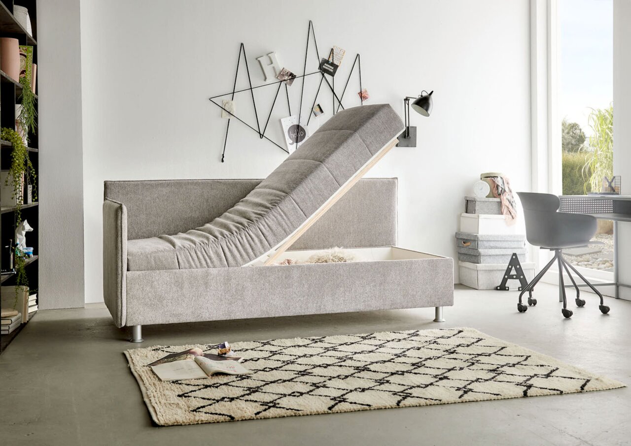 Graue Polsterliege mit hochgeklappter Matratze, seitliche Perspektive, zeigt Stauraum darunter, modernes Design in einem hellen Raum.