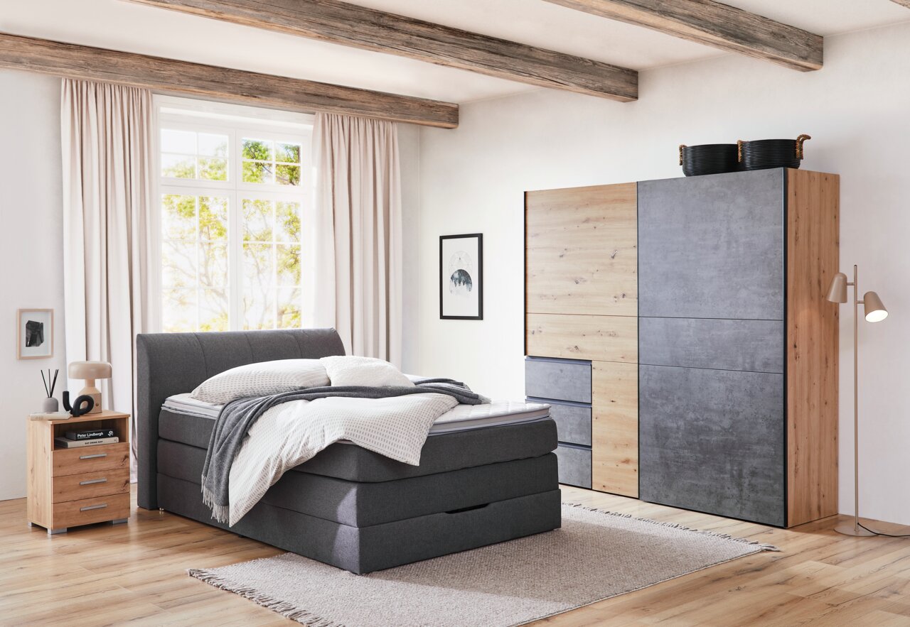 Moderner Schwebetürenschrank in Holz- und Betonoptik, seitliche Perspektive im Schlafzimmer mit Bett und Nachttisch.