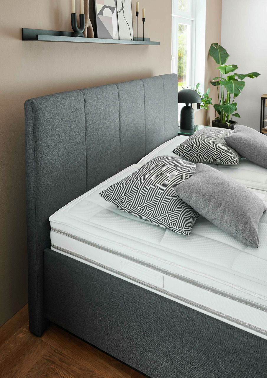 Seitliche Perspektive auf ein modernes graues Polsterbett mit hohem Kopfteil, dekoriert mit gemusterten Kissen und umgeben von stilvollen Accessoires.