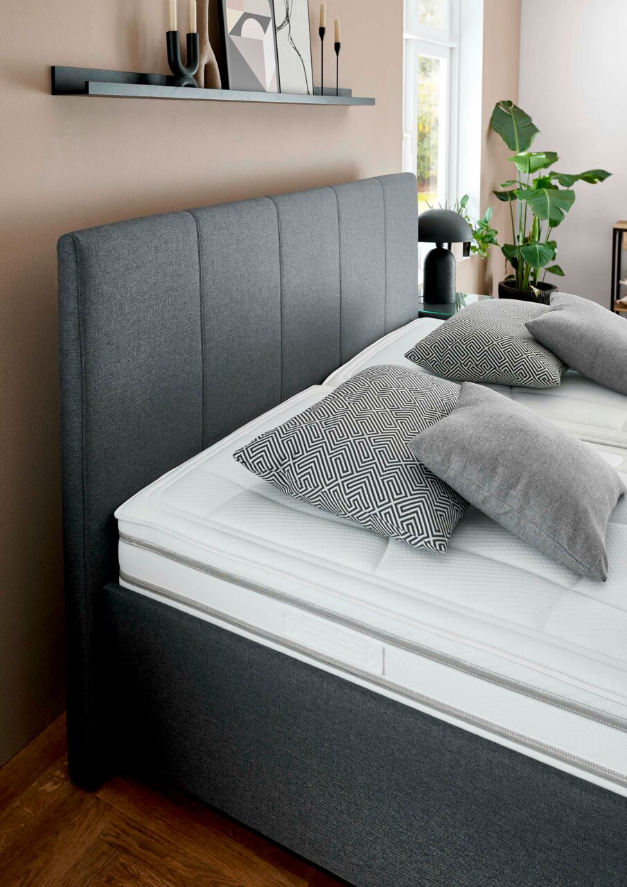 Seitliche Perspektive auf ein modernes graues Polsterbett mit hohem Kopfteil, dekoriert mit gemusterten Kissen und umgeben von stilvollen Accessoires.