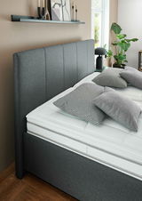 Seitliche Perspektive auf ein modernes graues Polsterbett mit hohem Kopfteil, dekoriert mit gemusterten Kissen und umgeben von stilvollen Accessoires.