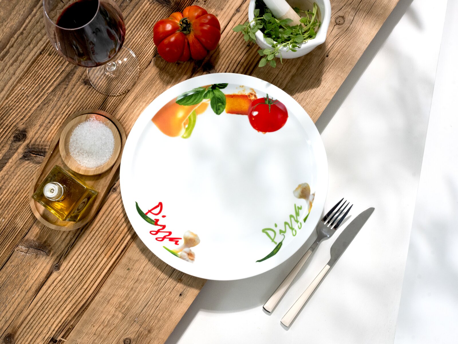 Runder weißer Pizzateller mit farbigem Gemüse-Dekor und „Pizza“-Schriftzug, auf rustikalem Holztisch neben Besteck, Wein, Salz, Olivenöl und Kräutern, fotografiert aus der Vogelperspektive (Draufsicht).