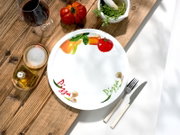 Runder weißer Pizzateller mit farbigem Gemüse-Dekor und „Pizza“-Schriftzug, auf rustikalem Holztisch neben Besteck, Wein, Salz, Olivenöl und Kräutern, fotografiert aus der Vogelperspektive (Draufsicht).