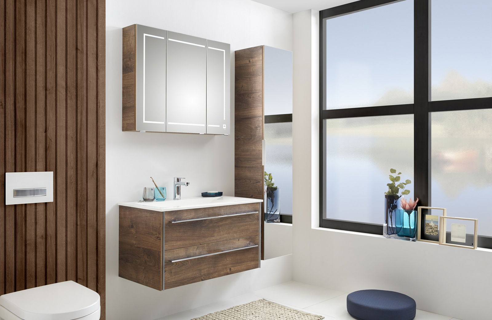 Moderne Badkombination mit Holzoptik, bestehend aus einem Waschtisch mit zwei Schubladen, einem beleuchteten Spiegelschrank und einem hohen Schrank, seitliche Perspektive.