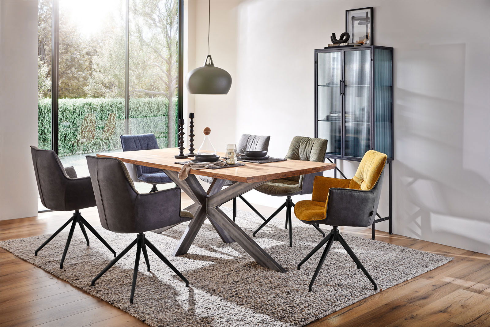 Esszimmer mit modernem Esstisch aus Holz und Metallgestell, umgeben von bunten Sesseln auf Teppich, mit Blick auf großen Fensterbereich.